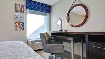 Quarto de hotel com cama, secretária e cadeira, espelho redondo e obras de arte na parede.