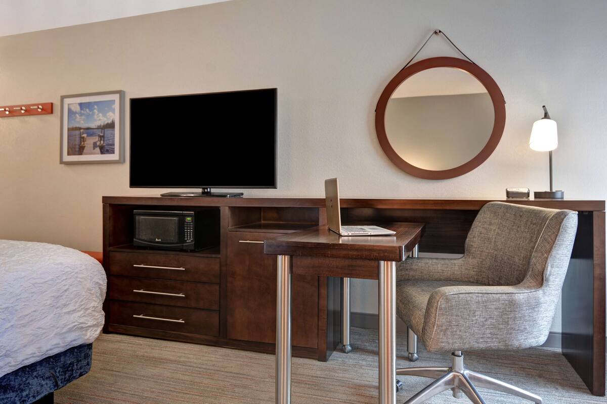 A hotel room with a bed, a desk with a laptop, a television, a microwave, a chair, a round mirror, and a lamp.