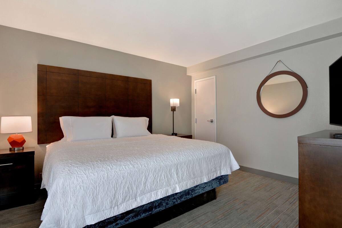 A hotel room with a large bed, a nightstand with a lamp, a dresser, a round mirror on the wall, and a television on the dresser.