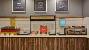 A breakfast buffet area with labeled food stations, including eggs, cereal, oatmeal, and bakery items.