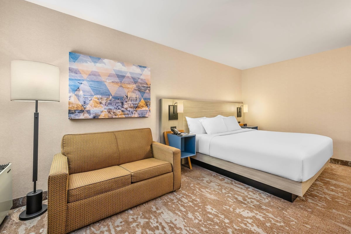 A hotel room with a bed, a sofa, a lamp, a painting, and a nightstand.