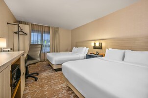 Quarto de hotel com duas camas, uma escrivaninha, uma cadeira, um abajur, uma mesa de cabeceira e uma janela com cortinas.