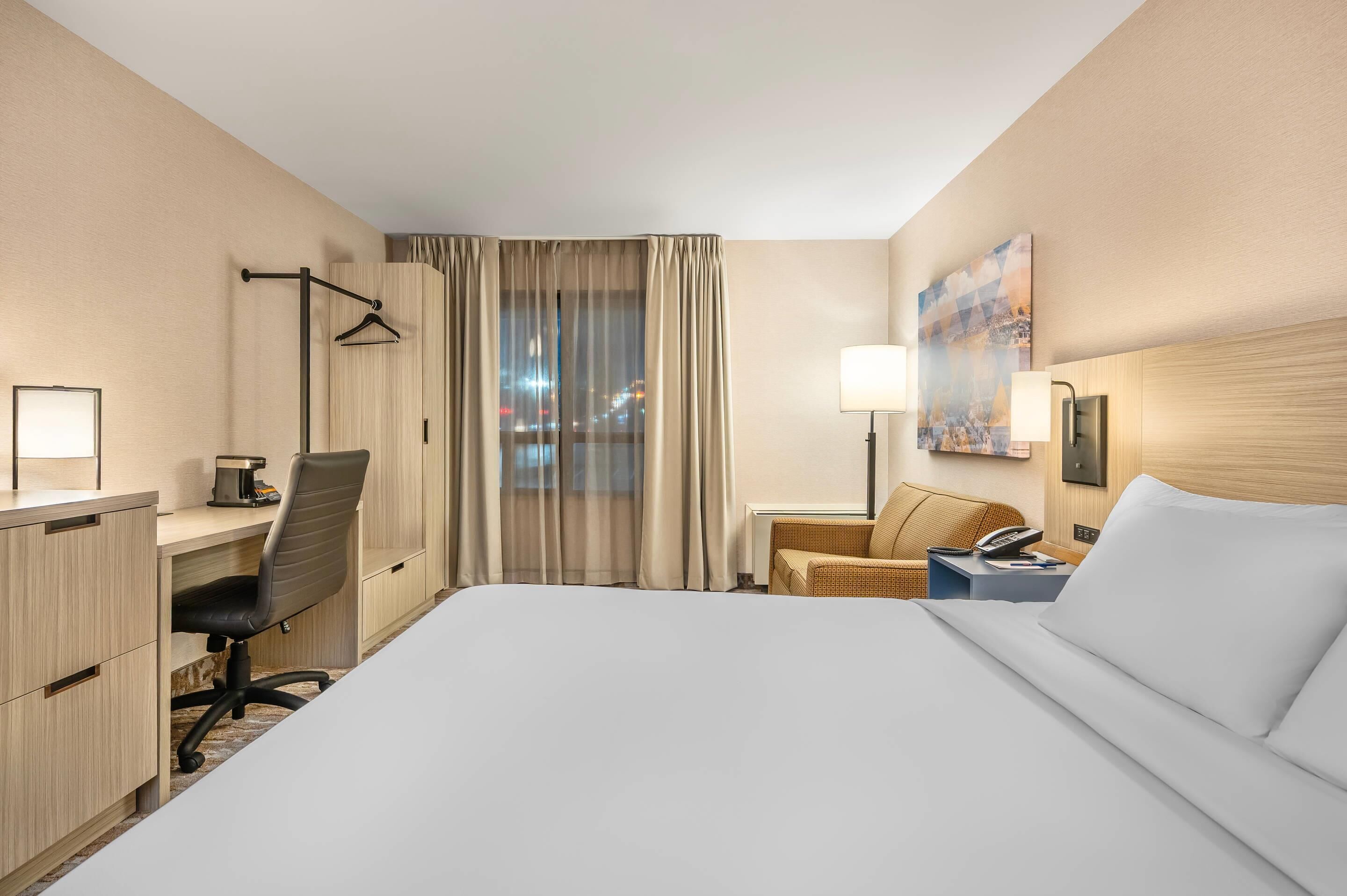 A hotel room with a large bed, a desk with a chair, a small table with a lamp, a phone, and a painting on the wall.