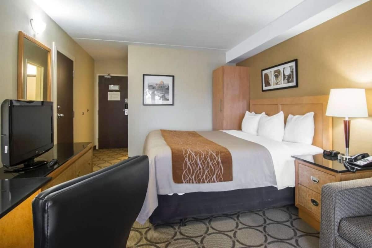 A hotel room with a bed, a desk with a television, a chair, a nightstand with a lamp, and a door.