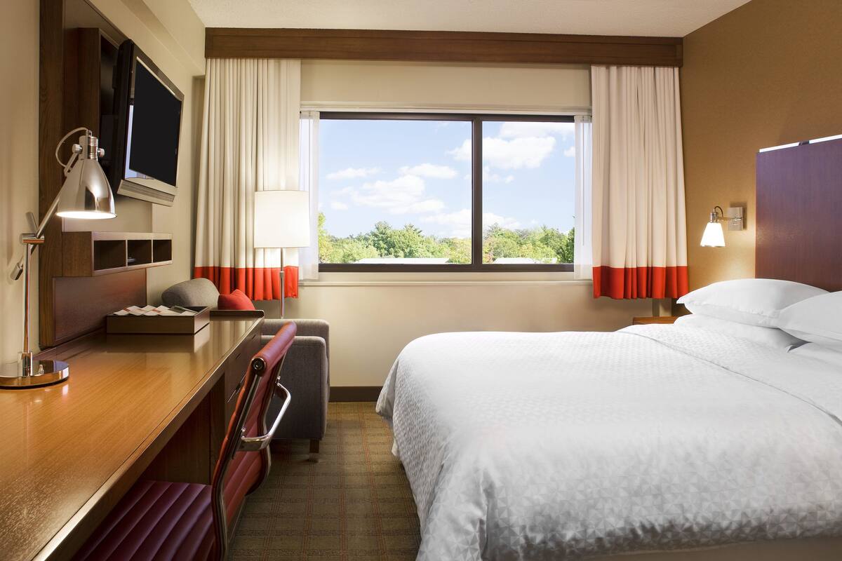 A hotel room with a bed, a desk, a chair, a TV, and a window with curtains.