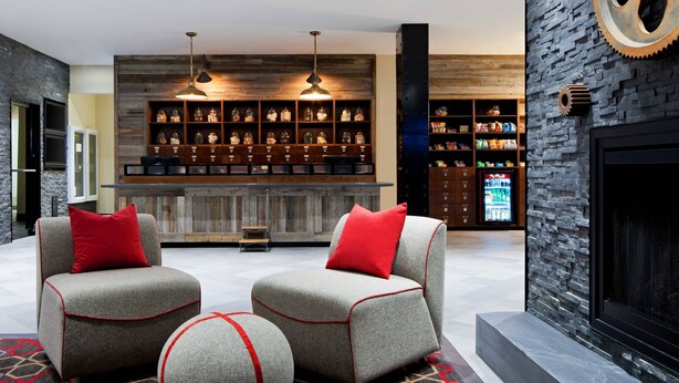 A modern lounge area with two grey armchairs with red accents, a patterned rug, a bar area with shelves of bottles, and a stone wall with a fireplace. - Four Points by Sheraton Edmundston Hotel & Conference Center (Edmundston)