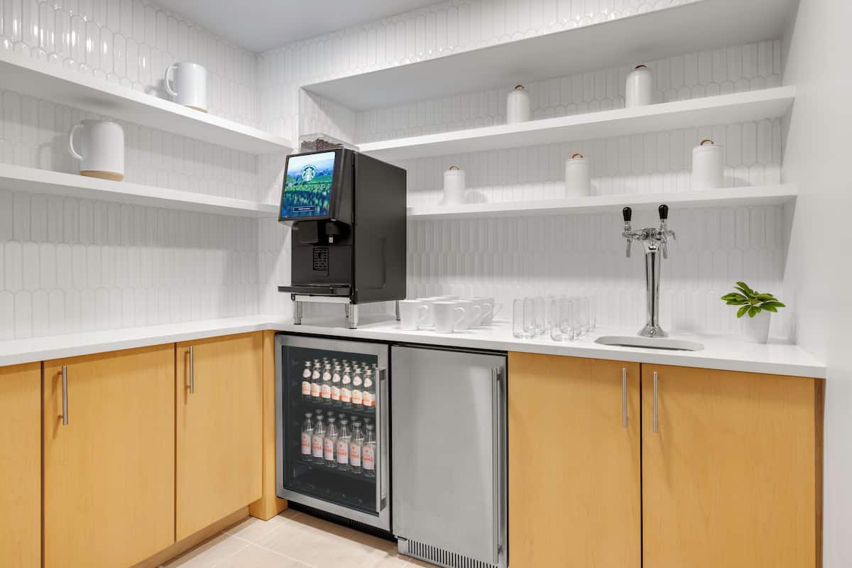 A modern kitchen with white cabinetry, a built-in coffee machine, a refrigerator, and a coffee station with cups and a plant.