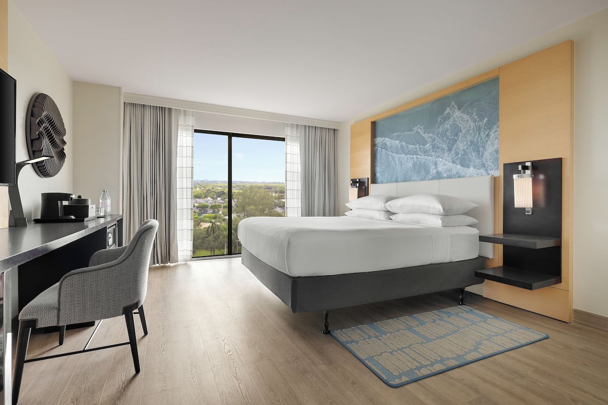 A modern hotel room with a large bed, a desk with a chair, a flat-screen TV, and a city view through the window.