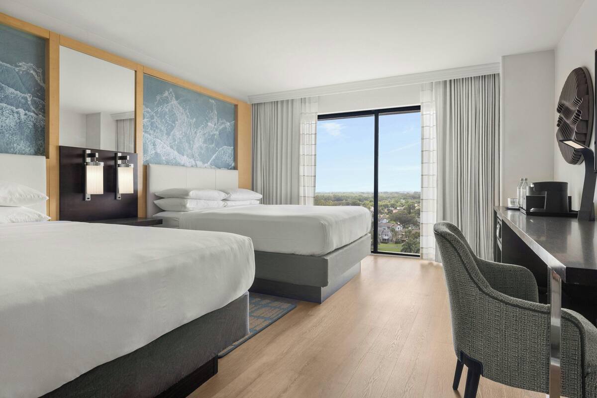 A modern hotel room with two beds, a desk, and a large window offering a city view.