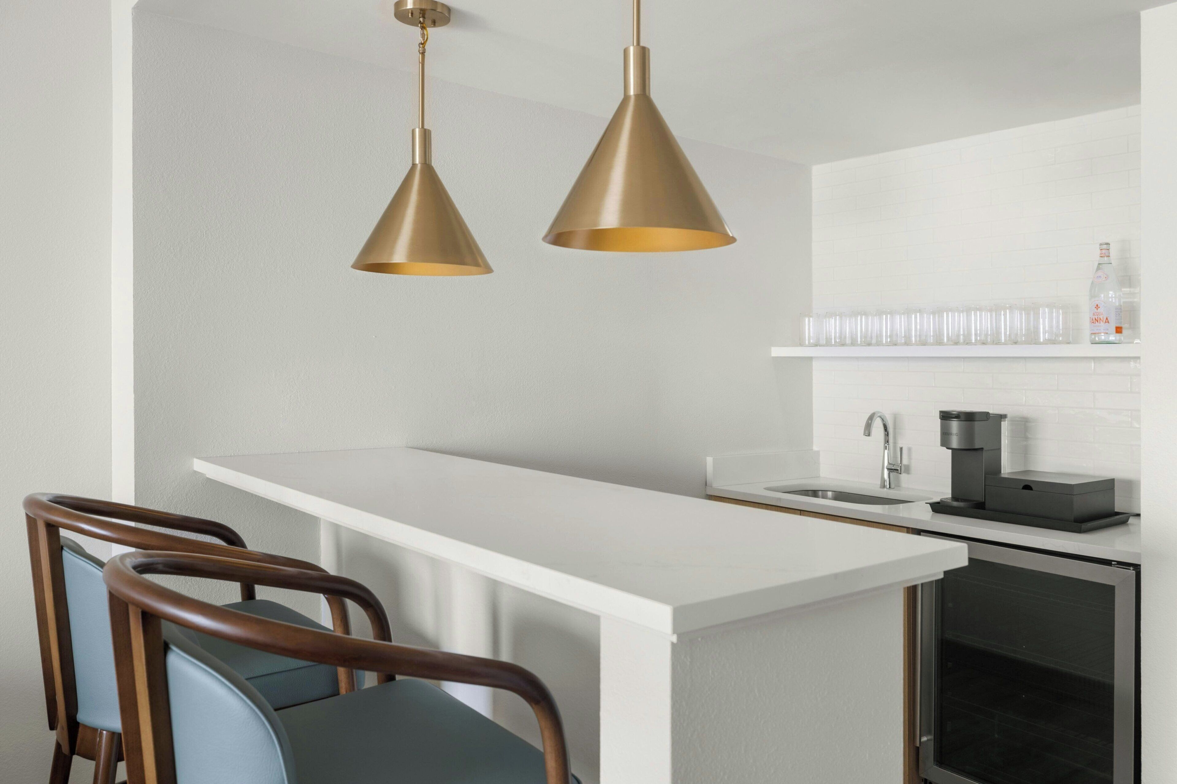 A modern kitchen with a white countertop, two pendant lights, a coffee machine, and a shelf with glasses.