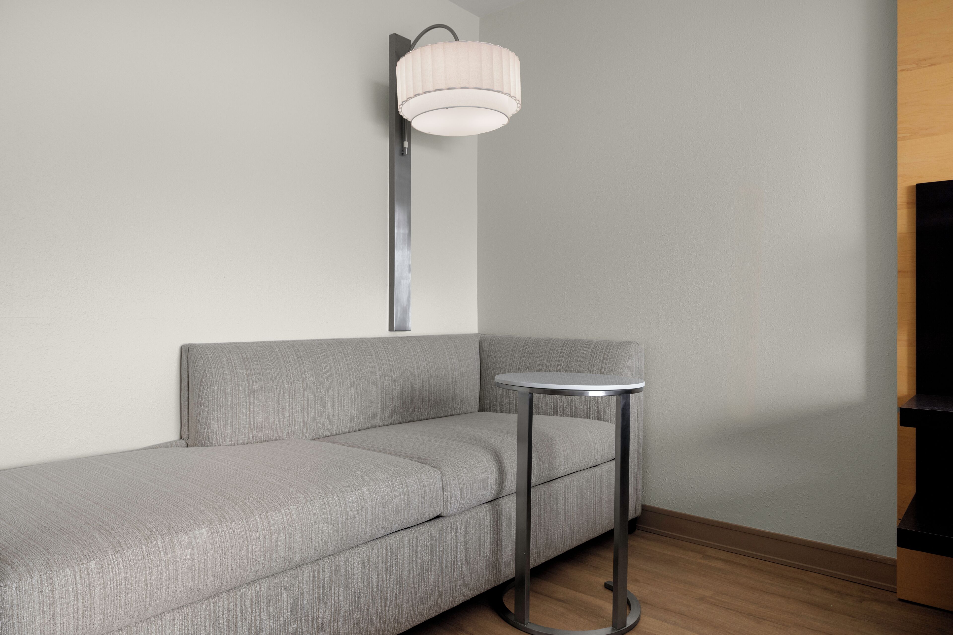 Un rincón de una habitación con un sofá gris, una mesa redonda de metal y una lámpara fijada a la pared.