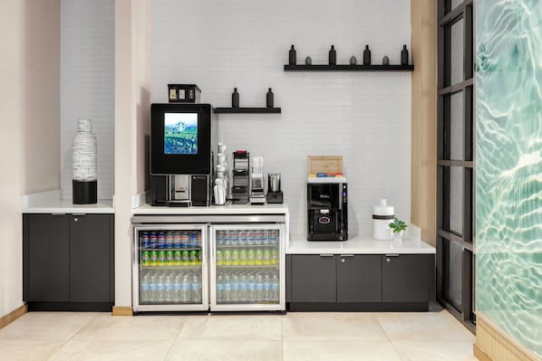 Un espace cuisine moderne comprenant une machine à café, un réfrigérateur rempli de boissons et un téléviseur diffusant un paysage.