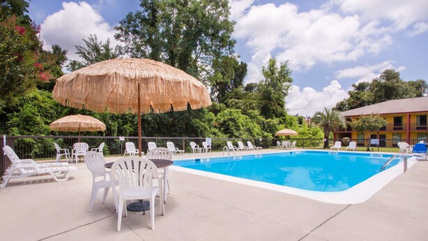 Una piscina con sillas de descanso y sombrillas de paja, rodeada de follaje y un edificio con balcones.