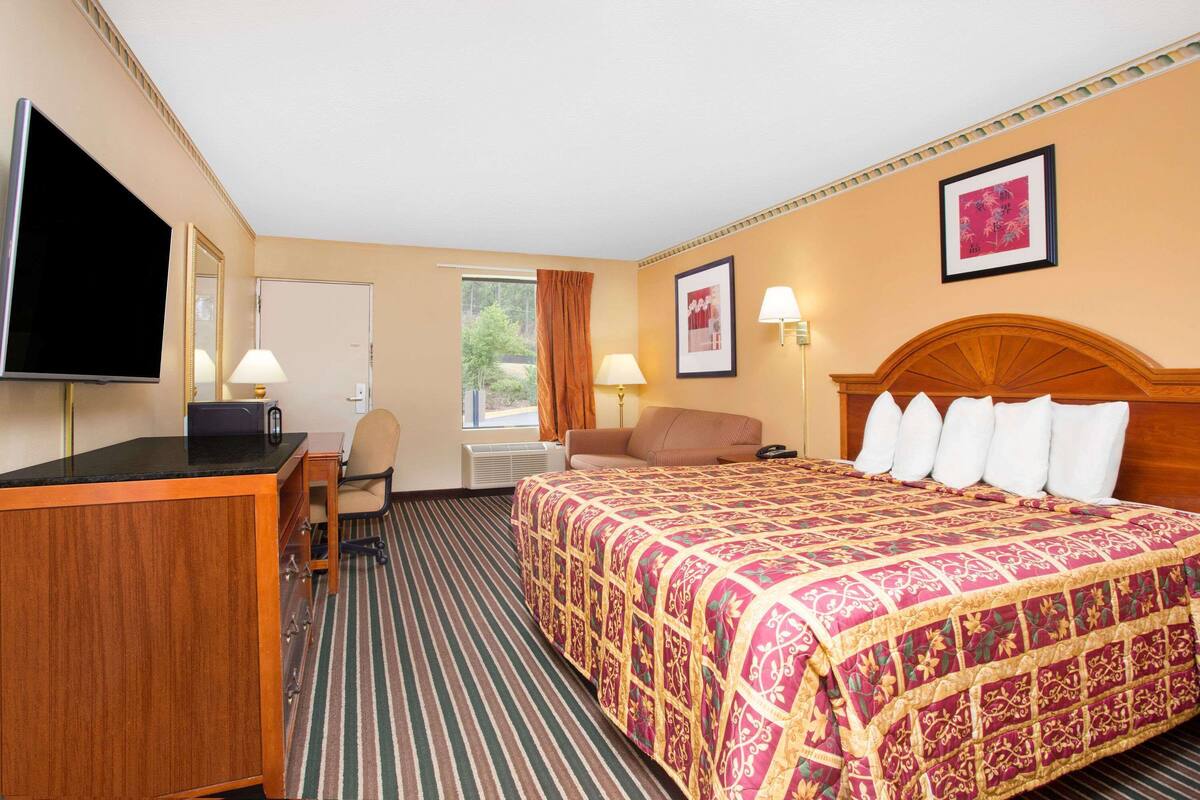 A hotel room with a large bed, a desk with a flat-screen TV, a chair, a window with curtains, and a framed picture on the wall.