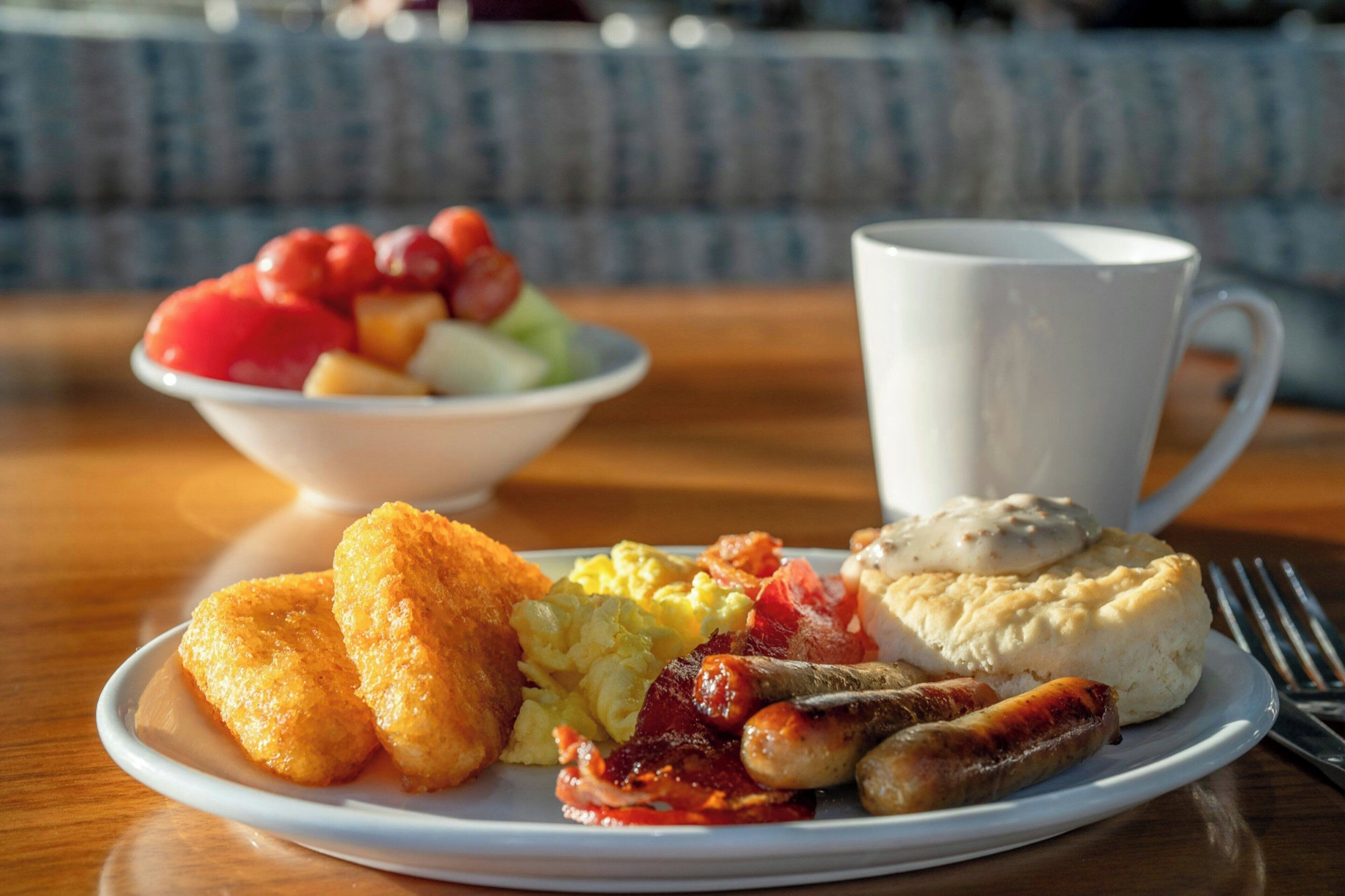 Une assiette de petit-déjeuner comprenant des œufs, du bacon, des saucisses, un biscuit et un bol de fruits.