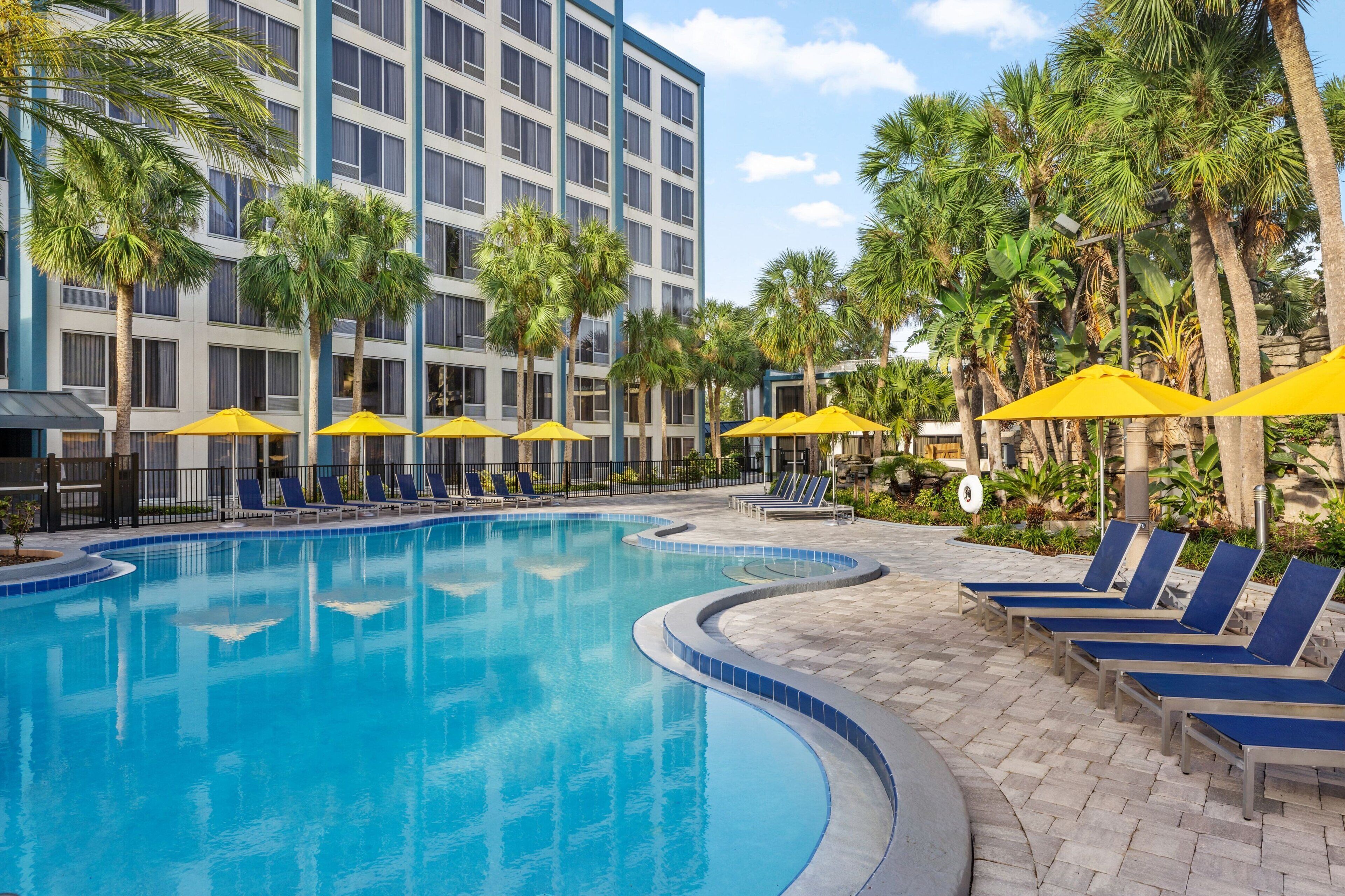 A swimming pool with blue lounge chairs and yellow umbrellas, surrounded by palm trees and a hotel building.