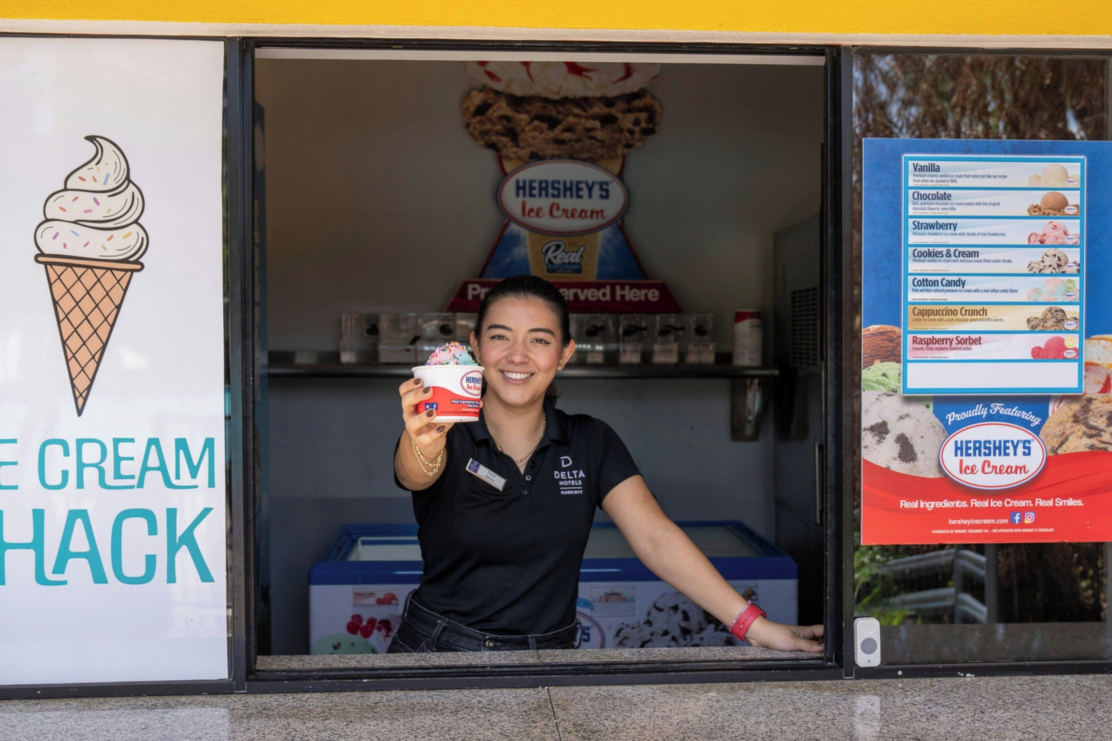 A person holding a Hershey's ice cream container in front of a store with a menu board displaying various ice cream flavors.