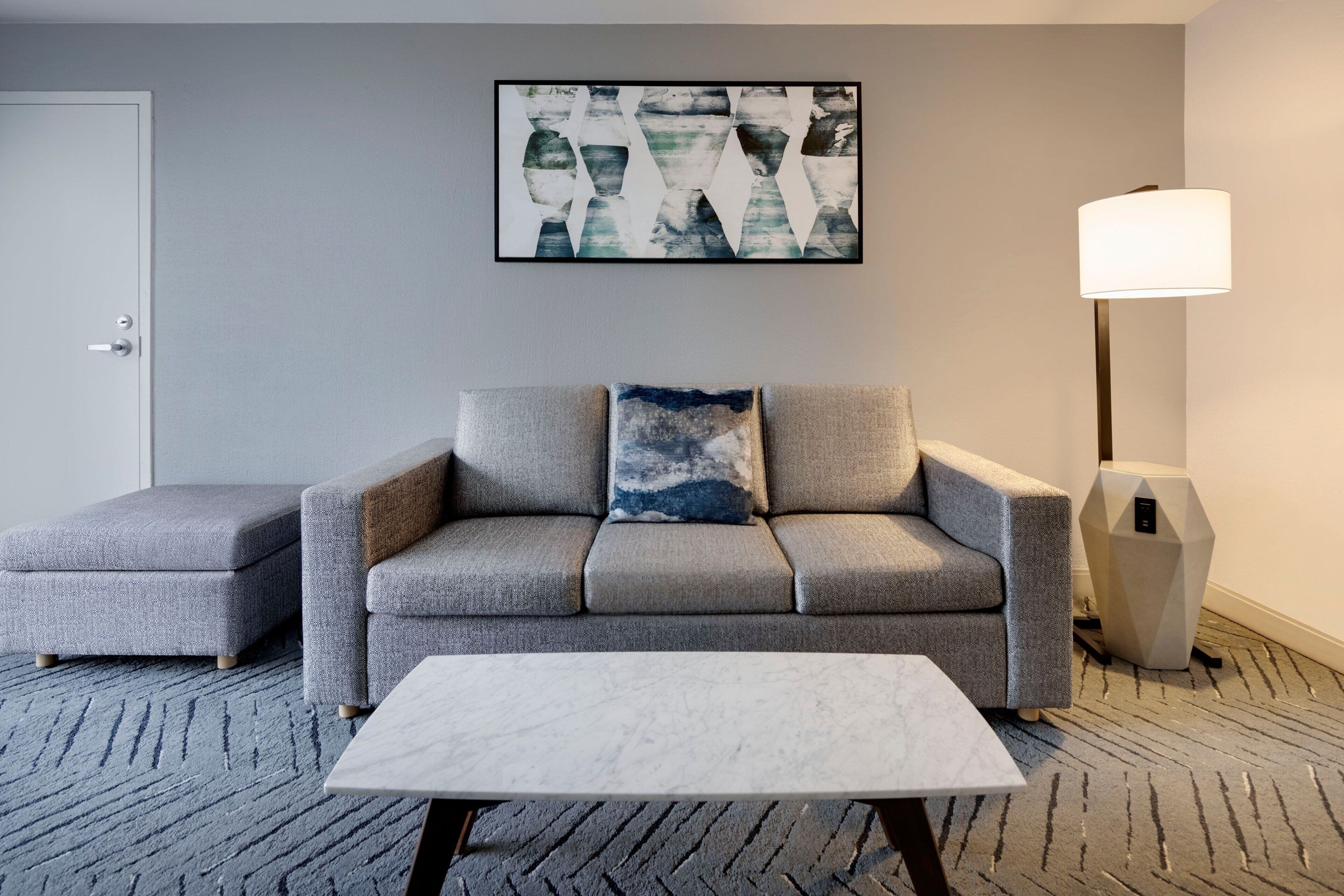 A modern hotel room with a grey sofa, a matching ottoman, a white coffee table, a floor lamp, and a large abstract painting on the wall.