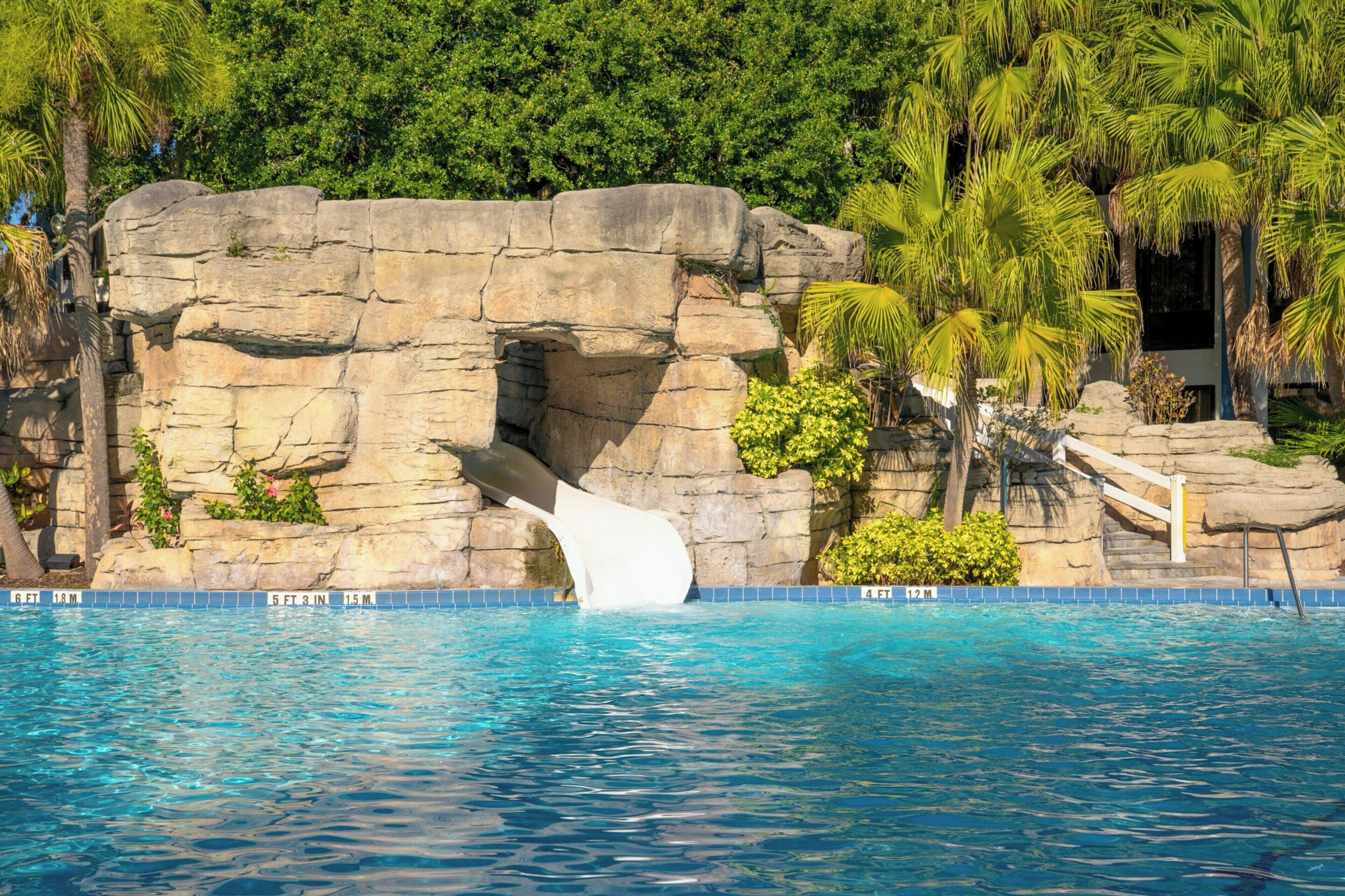 A pool with a rock waterfall feature, surrounded by lush greenery and palm trees.