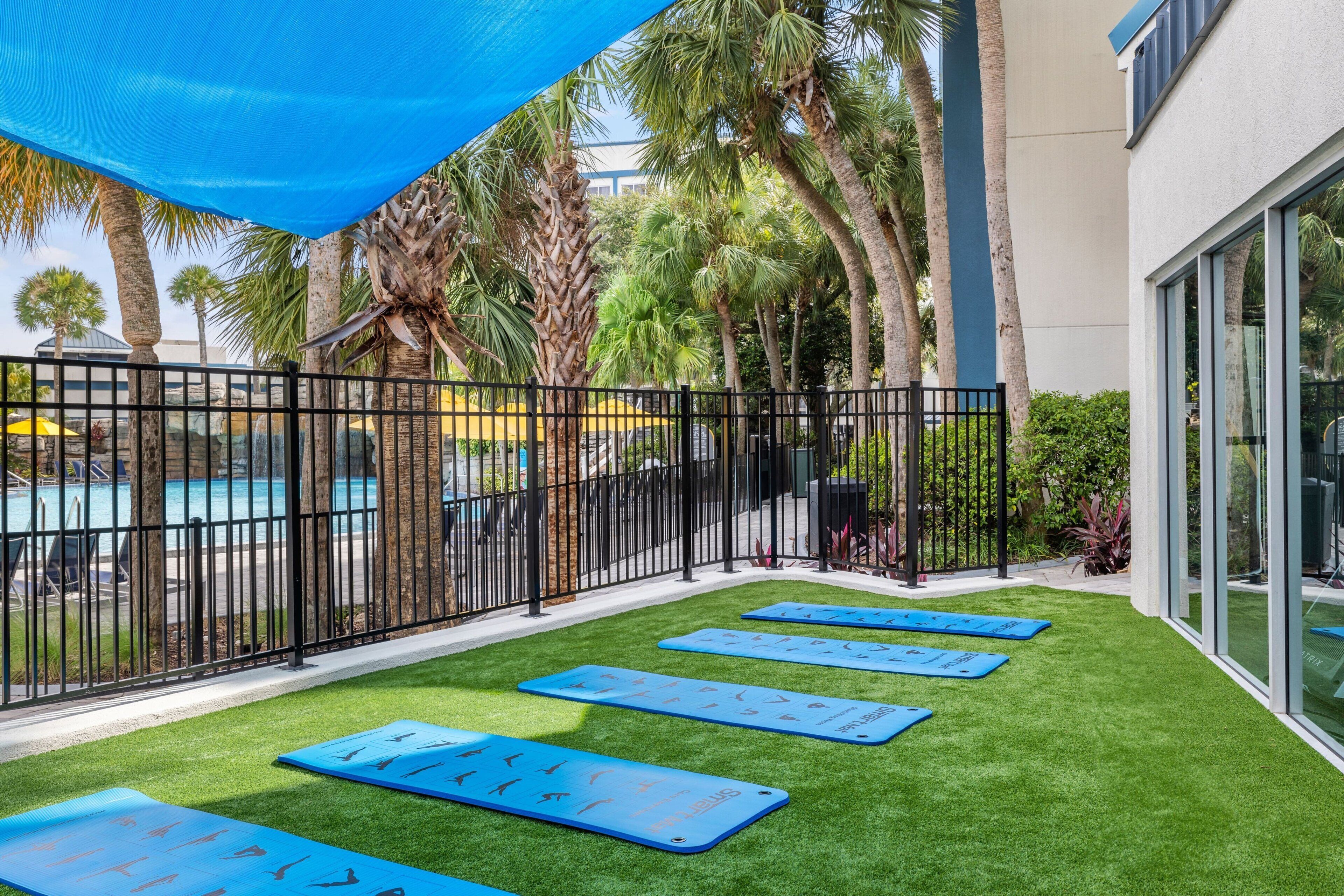 Un espace piscine avec des tapis bleus pour la pratique d’exercices, posés sur une pelouse artificielle, entouré d’une clôture en métal noir, de palmiers et d’un bâtiment doté de grandes fenêtres.