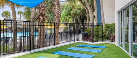 Un espace piscine avec des tapis bleus pour la pratique d’exercices, posés sur une pelouse artificielle, entouré d’une clôture en métal noir, de palmiers et d’un bâtiment doté de grandes fenêtres.