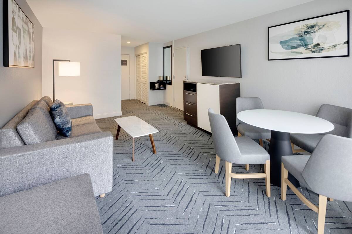 A modern hotel room with a grey sofa, a small white table, grey chairs, a round table, a flat-screen TV, and a herringbone-patterned carpet.