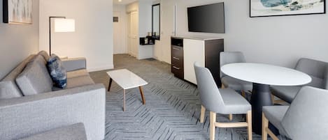 A modern hotel room with a grey sofa, a small white table, grey chairs, a round table, a flat-screen TV, and a herringbone-patterned carpet.