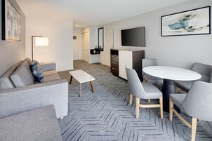 Une chambre d’hôtel moderne avec un canapé gris, une petite table blanche, des chaises grises, une table ronde, un téléviseur à écran plat et un tapis à motifs chevrons.