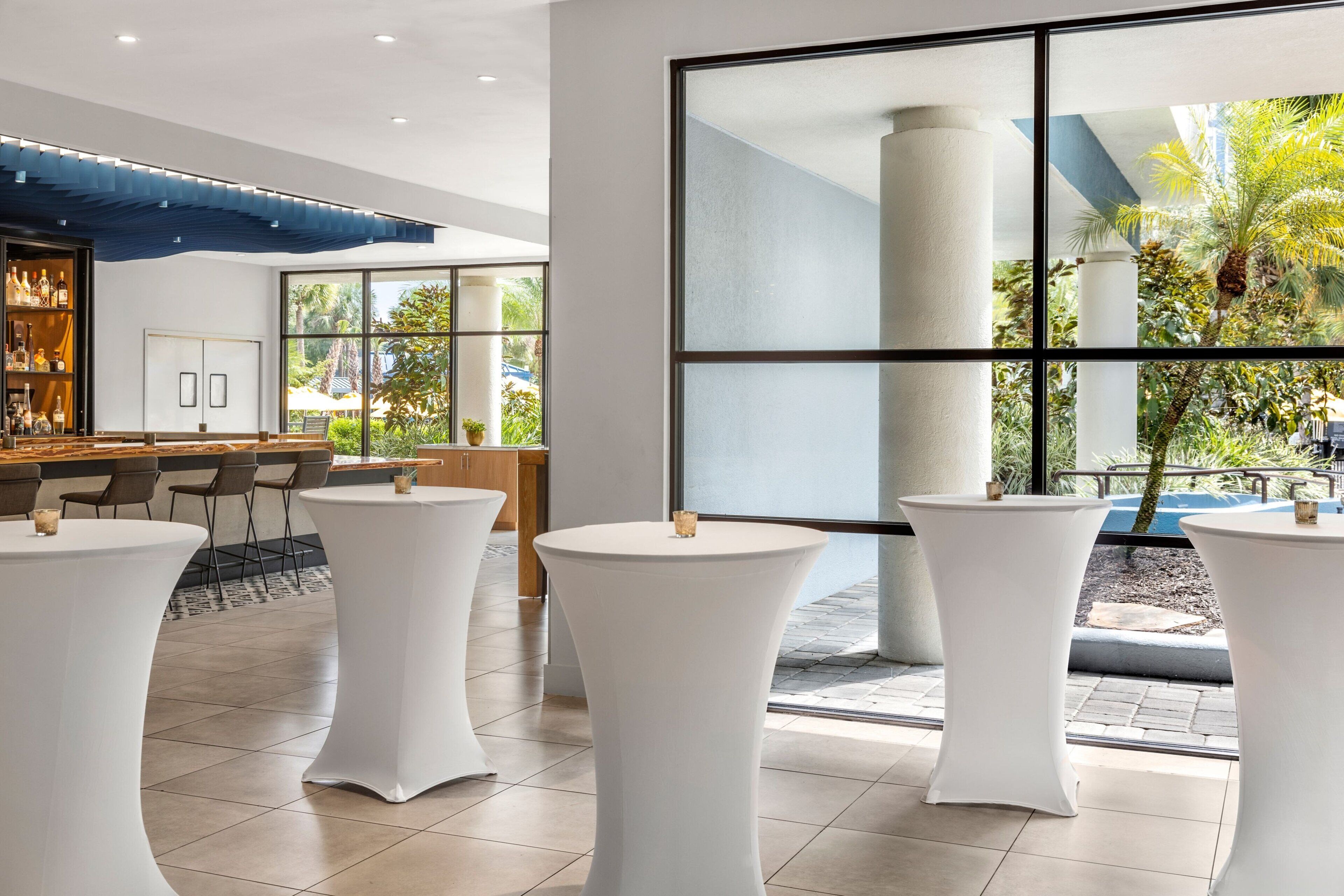 A modern hotel lobby with white round tables, a bar area, and large windows offering views of the outdoors.