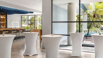 A modern hotel lobby with white round tables, a bar area, and large windows offering views of the outdoors.