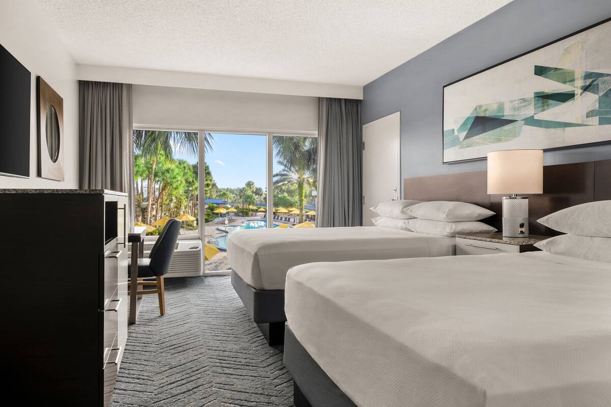 A hotel room with two beds, a desk, and a chair. There is a large window with a view of a pool and palm trees.