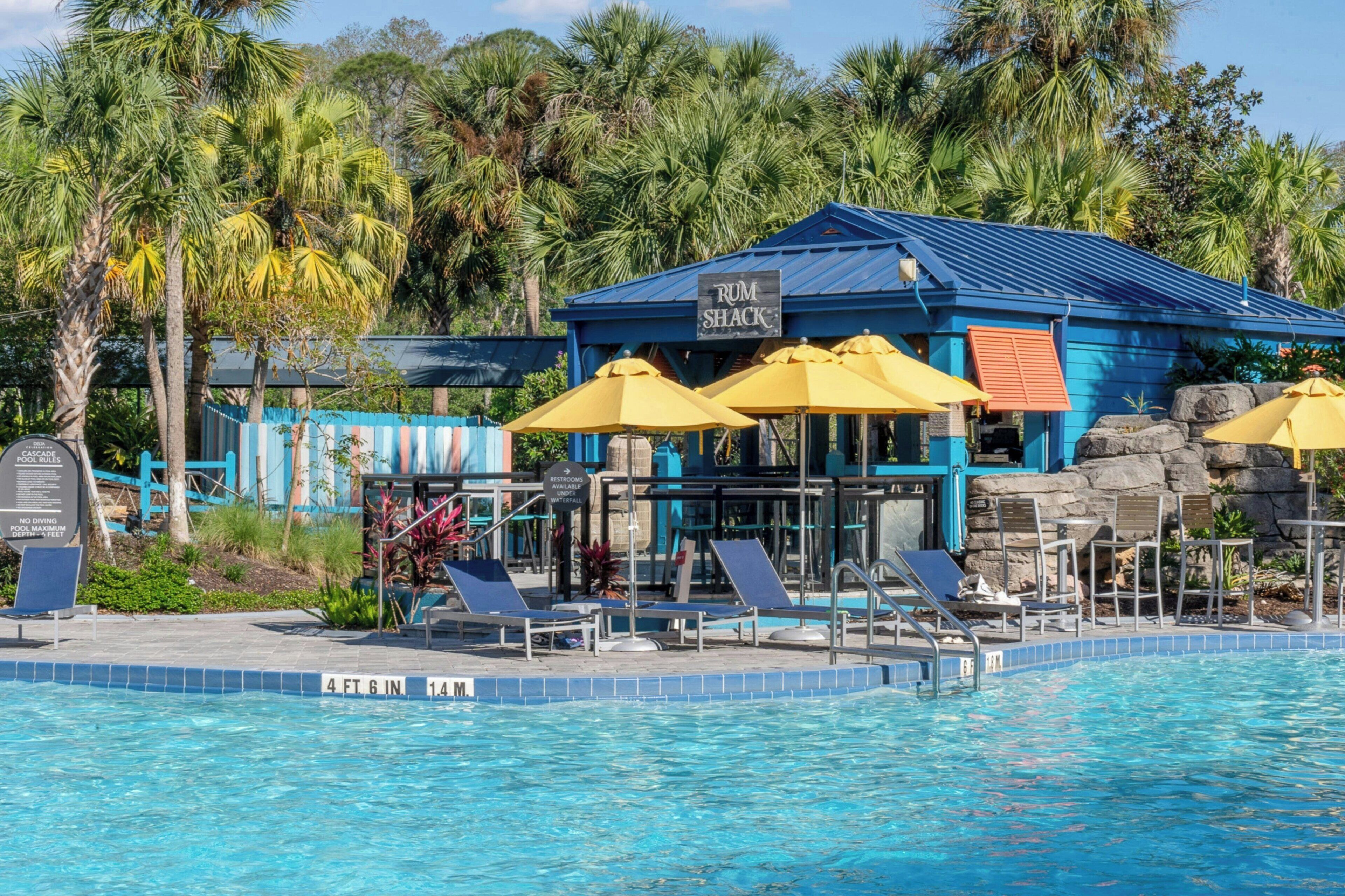 A pool area with lounge chairs and umbrellas, a rum shack, and tropical vegetation.