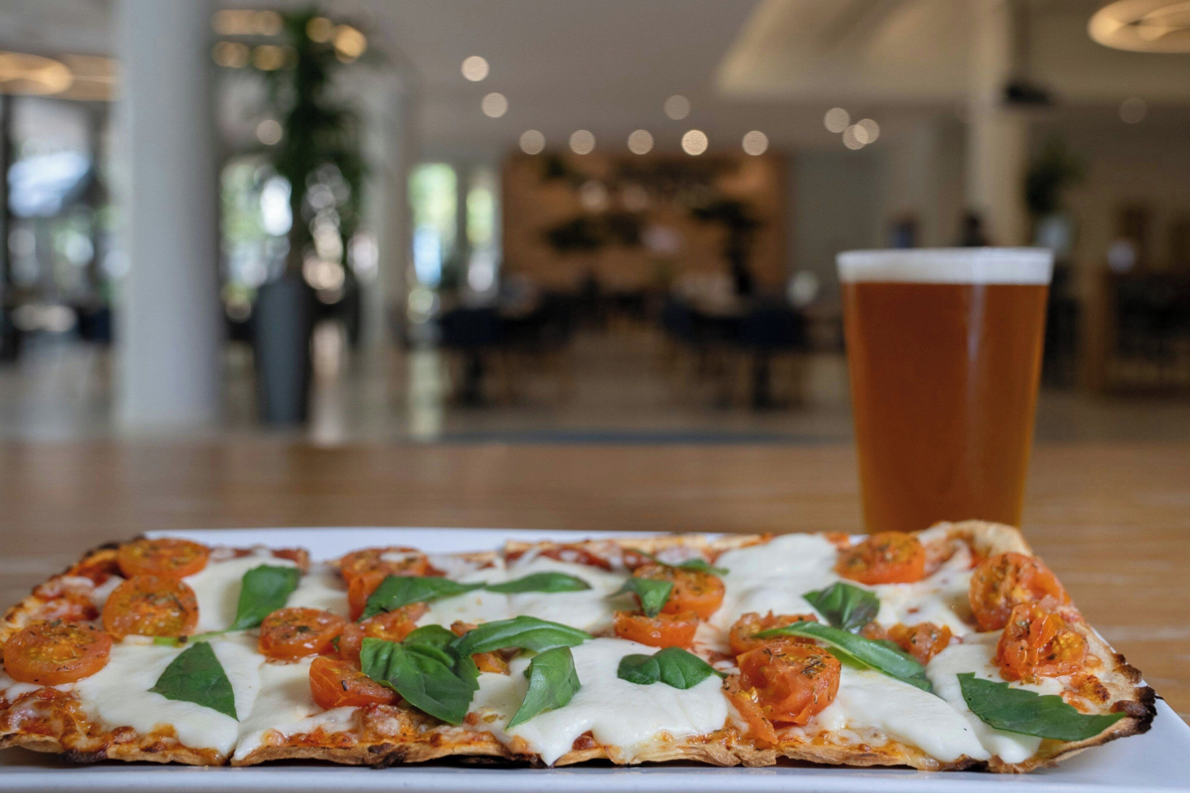 Pizza, jossa on tomaatteja ja basilikaa, valkoisella lautasella. Sen vieressä lasi olutta, ja taustalla on epätarkka kuva tilavasta sisätilasta.