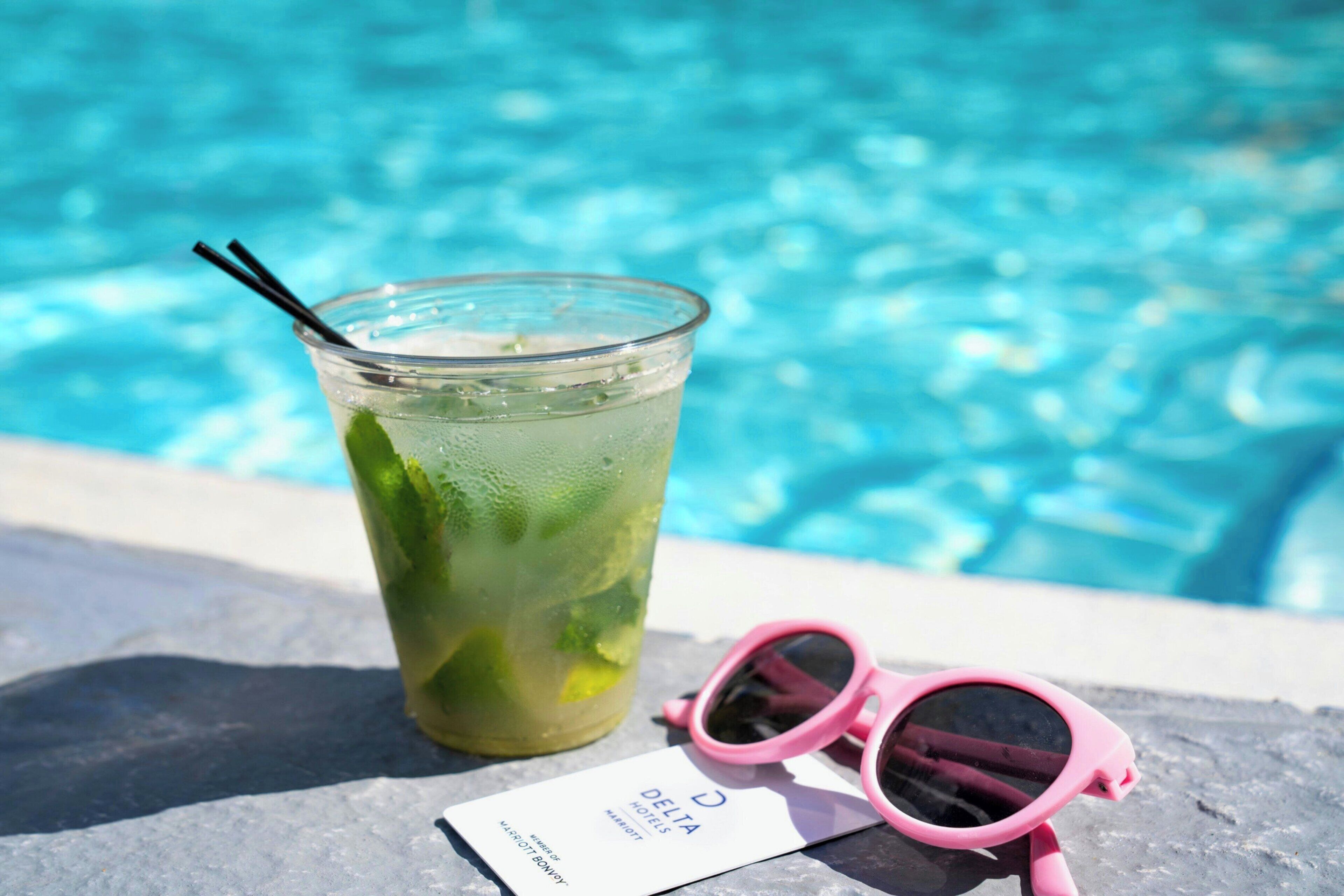Un verre transparent contenant une boisson verte, une paille, des lunettes de soleil roses et une carte de visite sur laquelle on peut lire « DELTA HOTELS », le tout posé sur une surface en pierre, près d’une piscine.