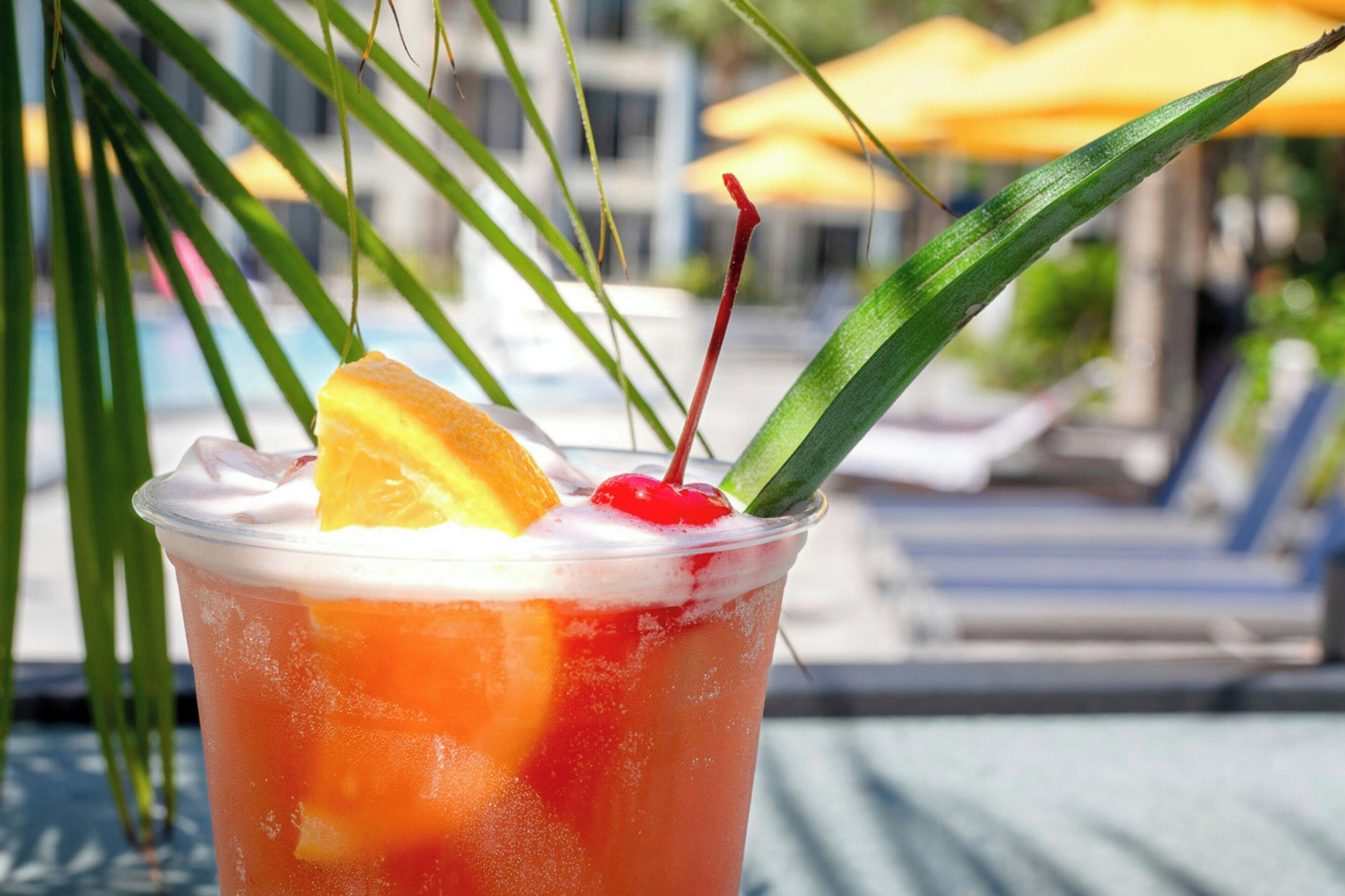 A tropical drink with a cherry and orange slice, served in a clear plastic cup, with a poolside background.