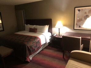 A hotel room with a bed, a desk, a chair, a lamp, and a framed picture on the wall.
