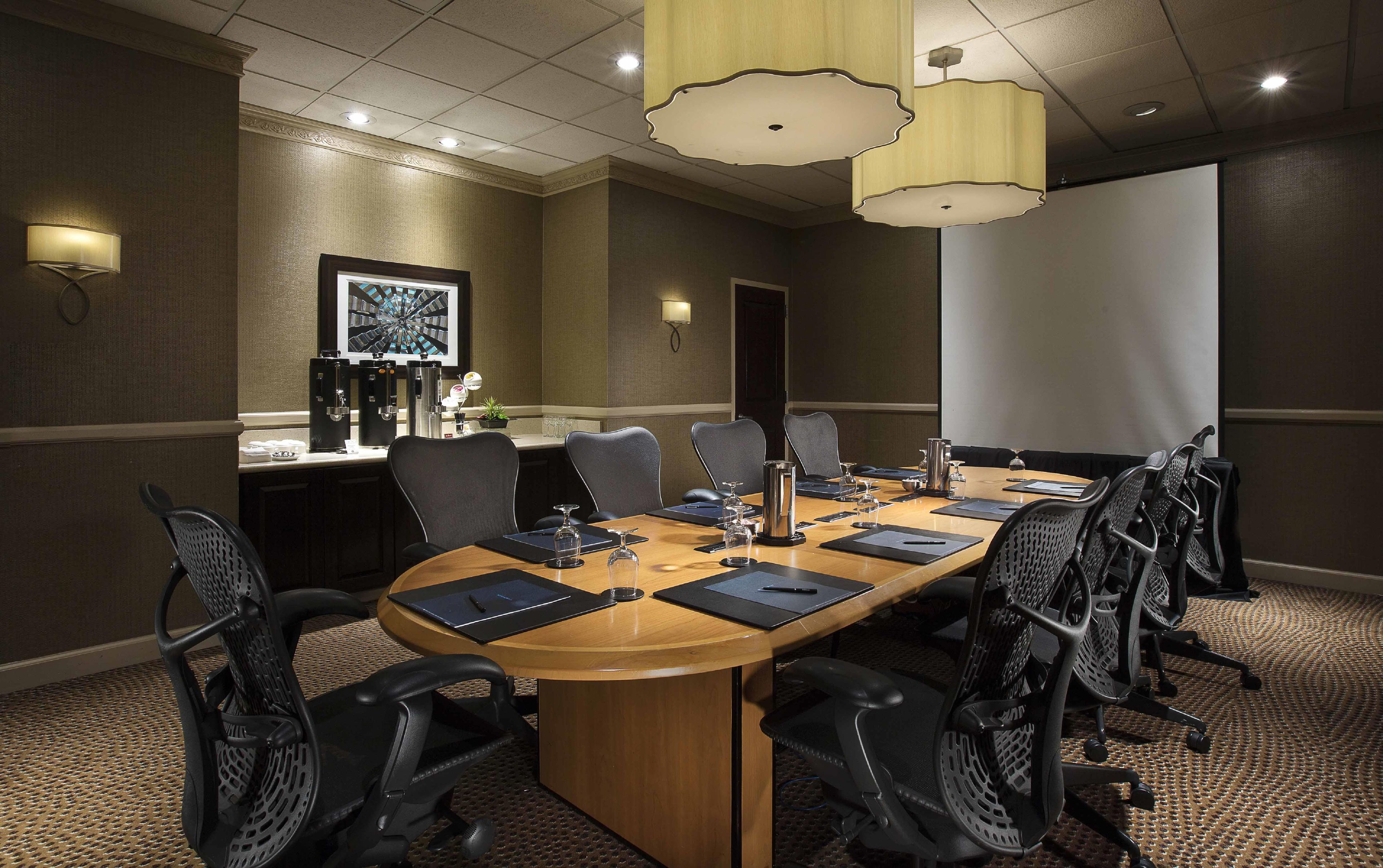 A conference room with a long table, multiple chairs, a projector screen, and a coffee station.
