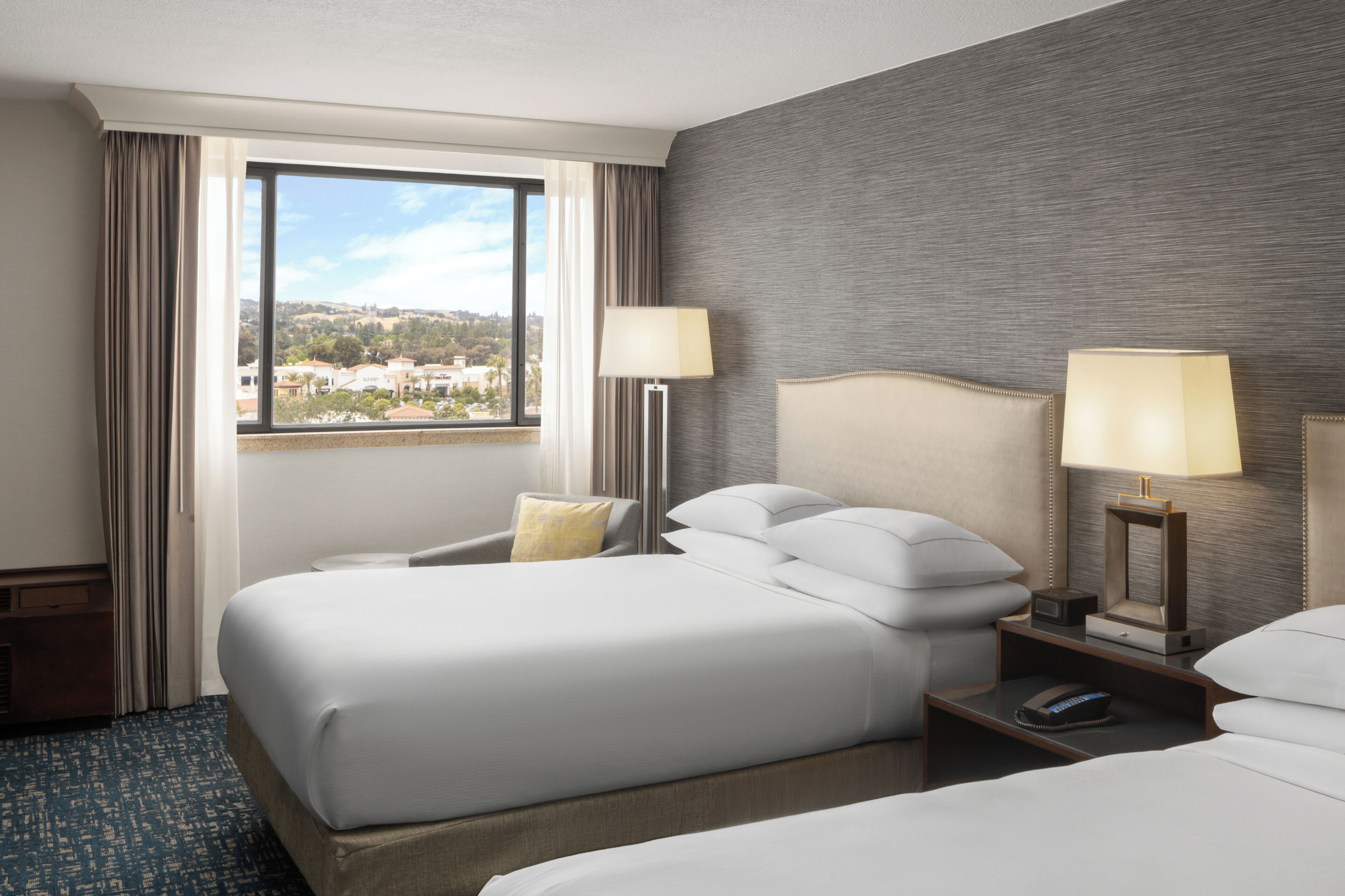 A hotel room with two beds, a large window, a chair, and a nightstand with lamps.