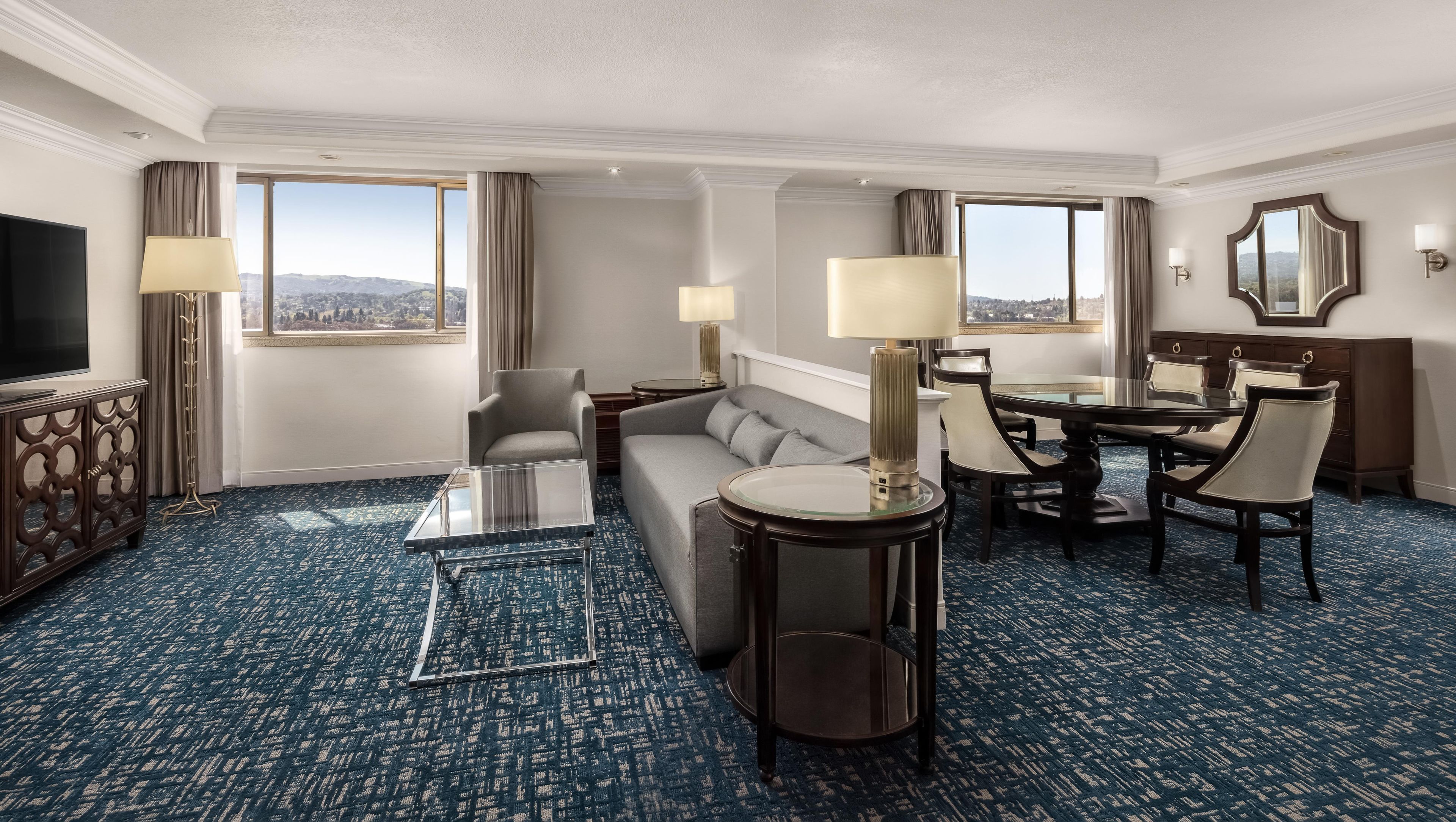 A hotel room with a sofa, armchair, dining table, chairs, and a TV.