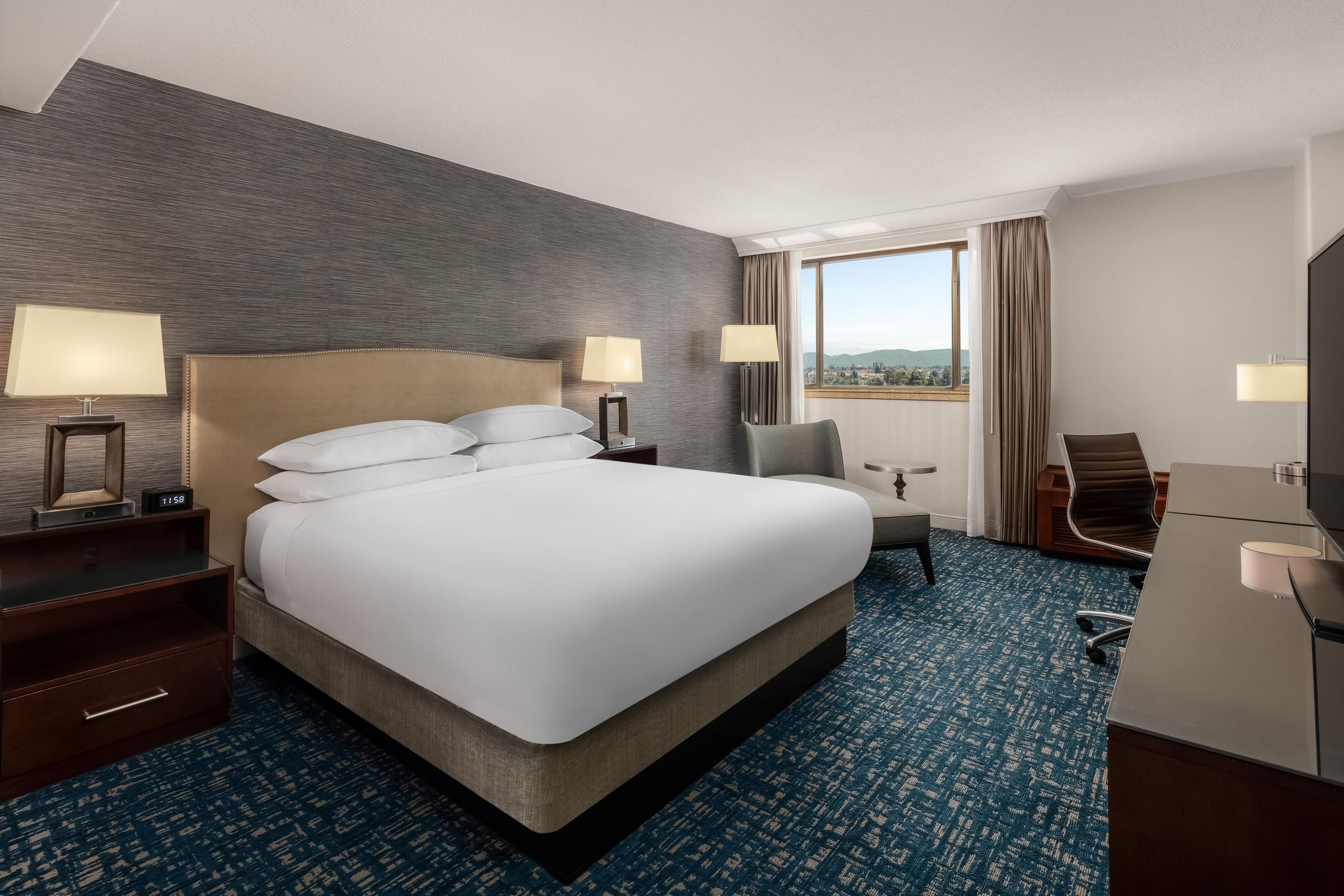 A hotel room with a large bed, two bedside tables with lamps, a desk with a chair, and a view of the outdoors through a window.