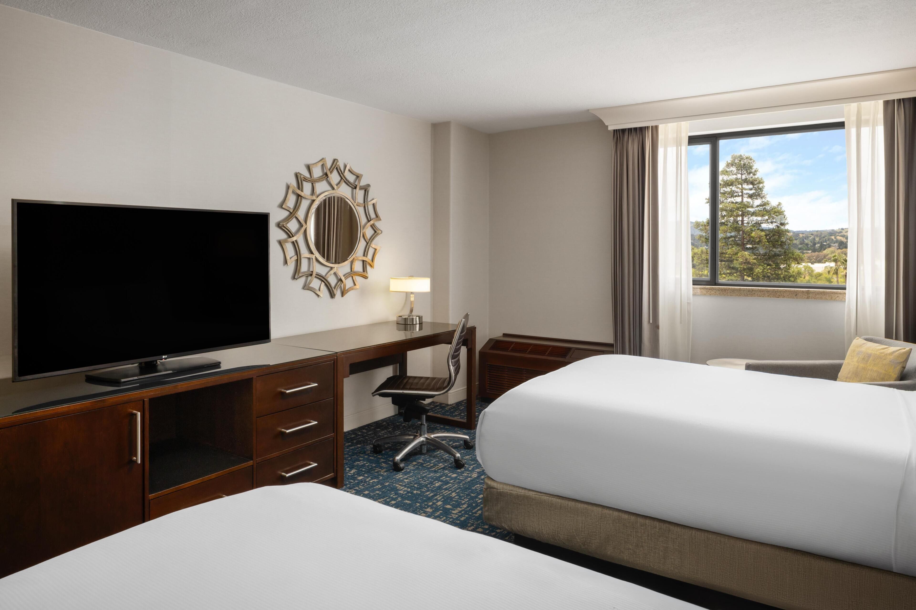 A hotel room with a TV, desk, two beds, a chair, and a window with a view.