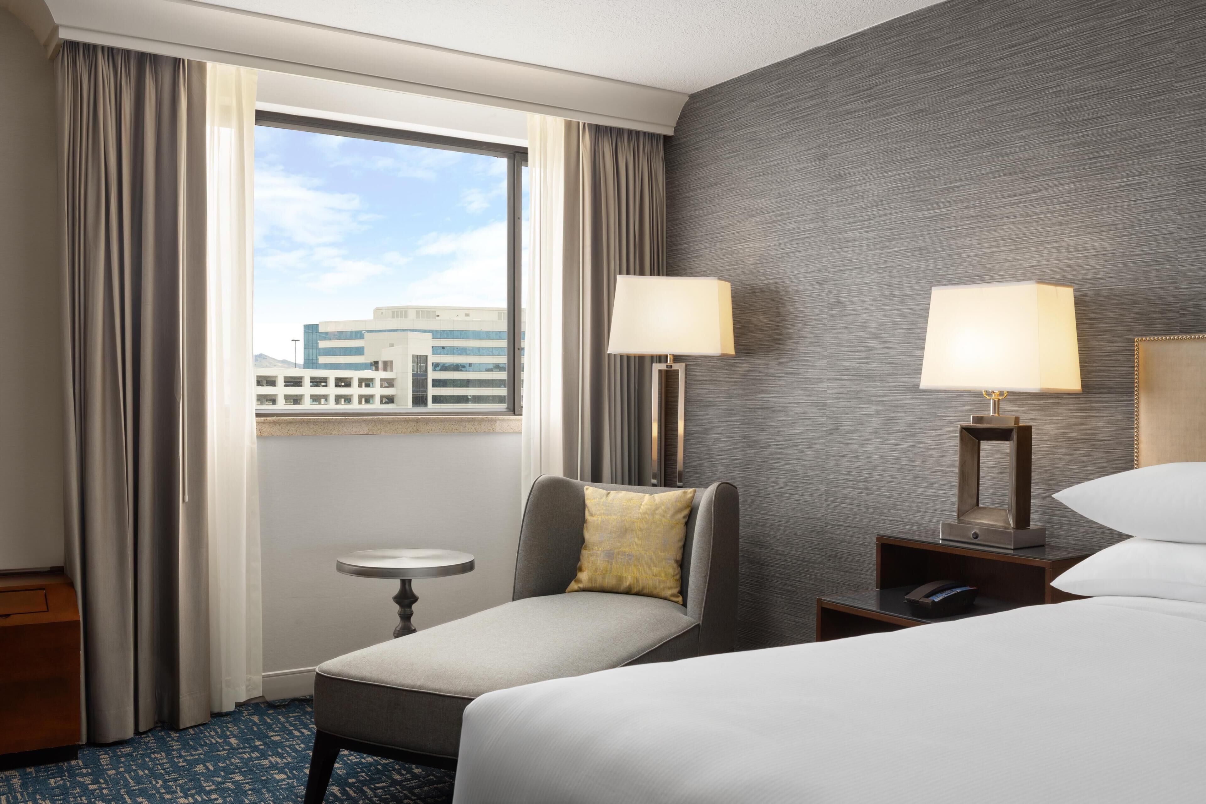Habitación de hotel con cama, una mesita, una silla, una lámpara y vistas a un edificio desde la ventana.