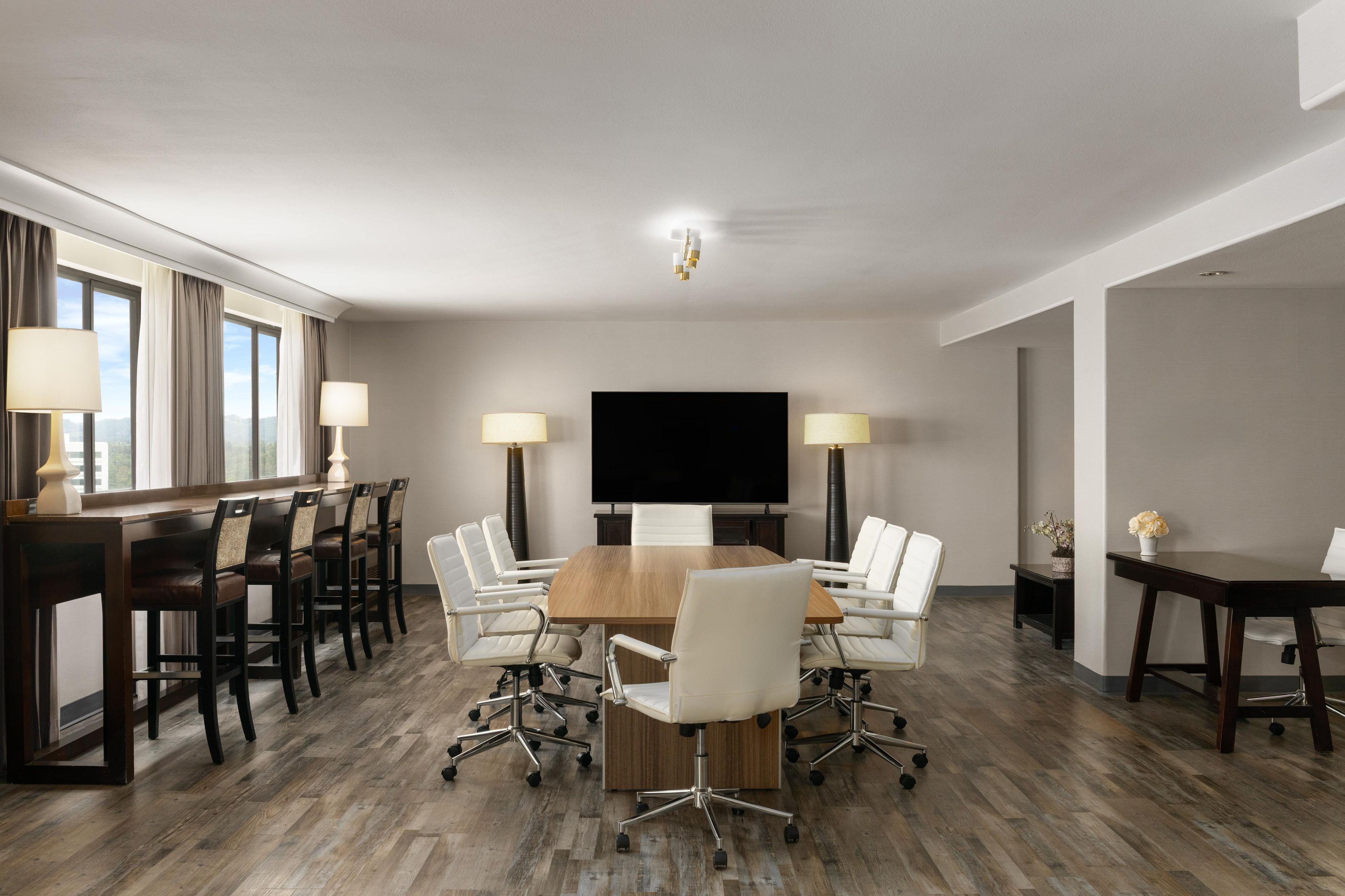 A conference room with a large table, white chairs, a flat-screen TV, and lamps.