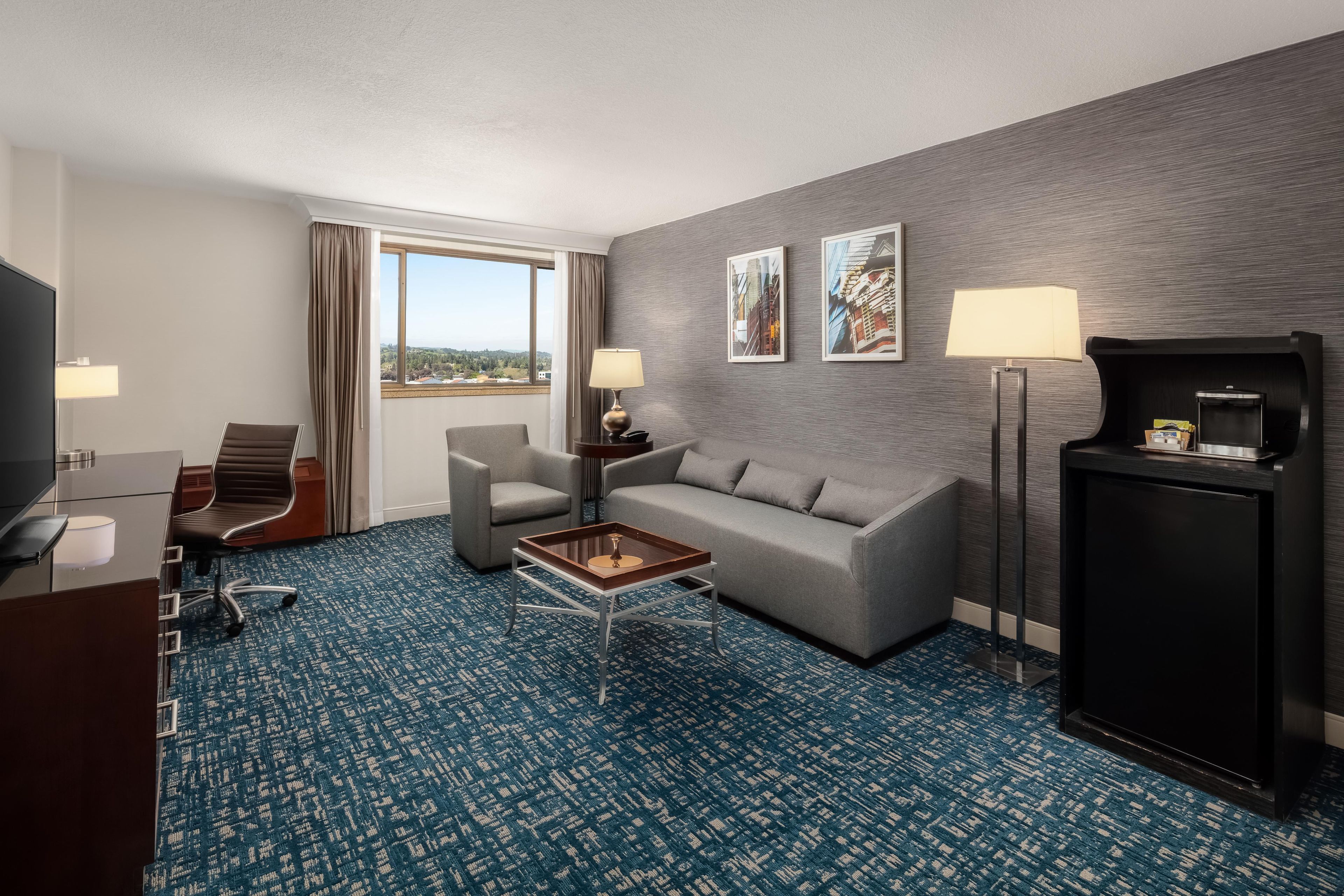 A hotel room with a grey sofa, a coffee table, a TV, a desk, and a window with curtains.