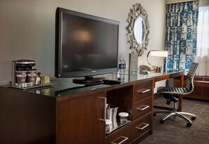 Habitación de hotel con televisor de pantalla plana, cafetera, minibar, escritorio con silla y espejo en la pared.