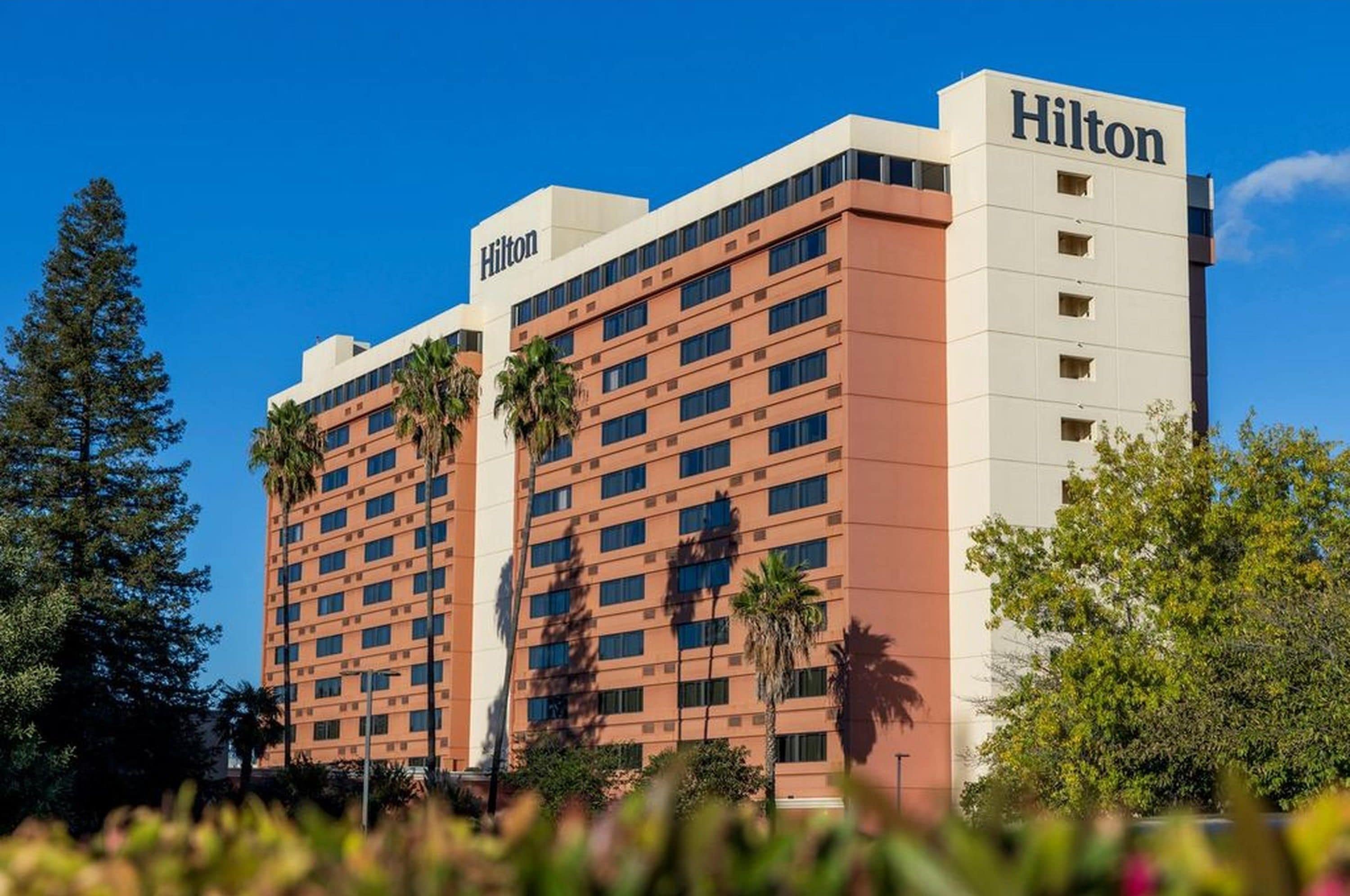 A Hilton hotel building with multiple floors, a prominent 'Hilton' sign on the top, and a clear blue sky in the background.