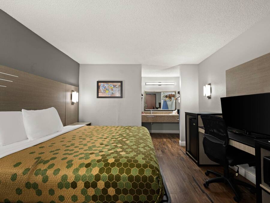 A hotel room with a bed, a desk with a computer, a TV, and a bathroom visible in the background.