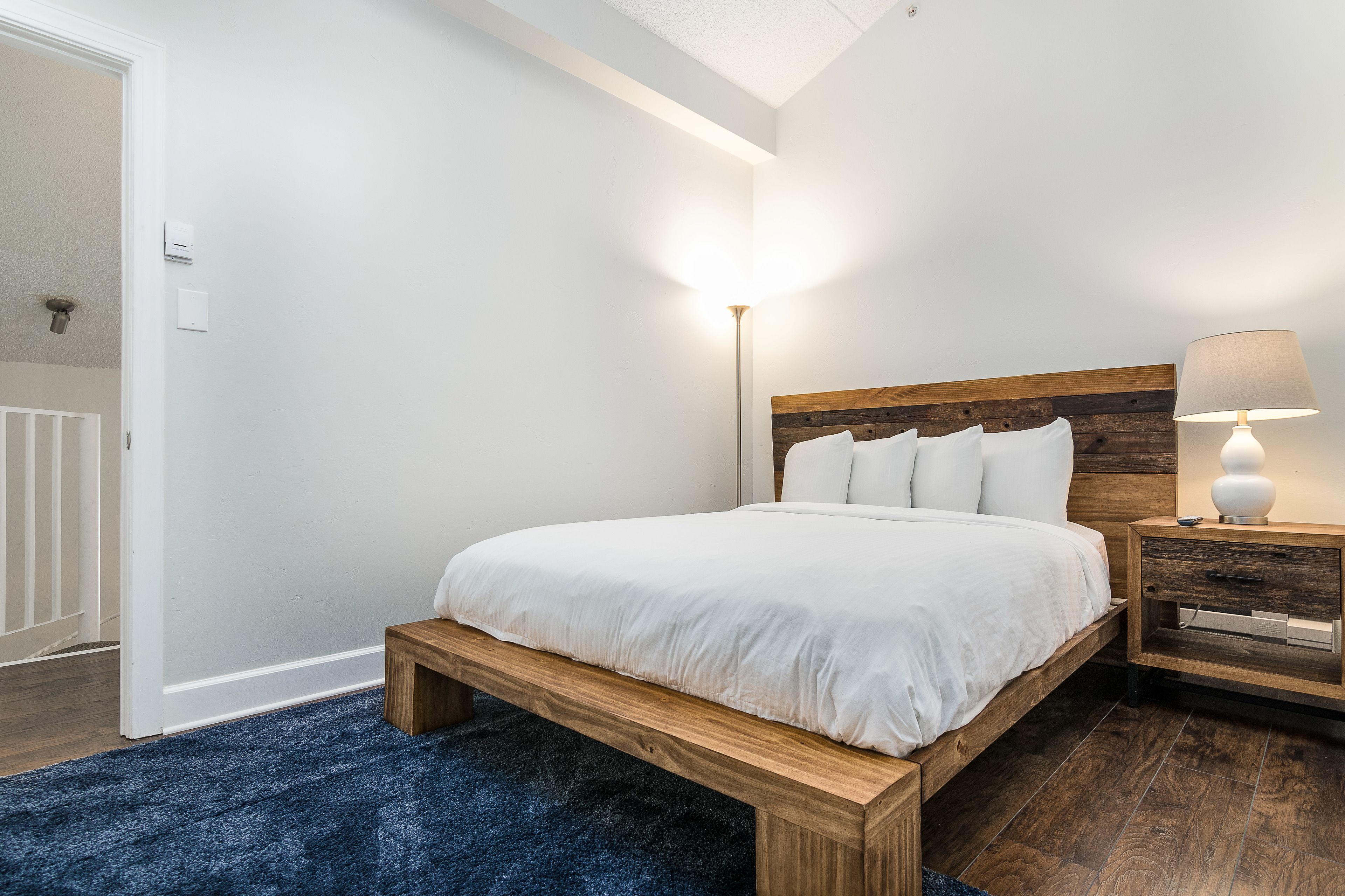 A bedroom with a wooden headboard, a white bedspread, a bedside table with a lamp, and a blue carpet.
