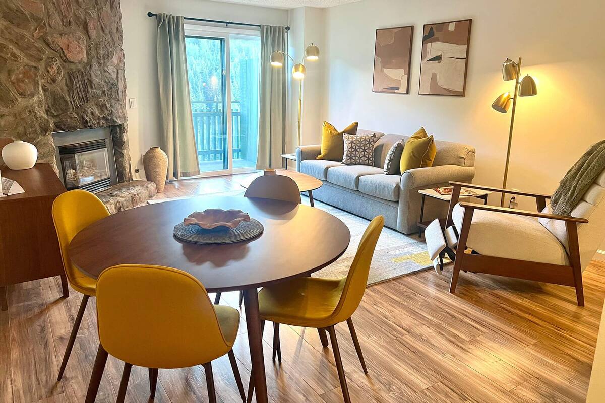 Una sala de estar moderna con chimenea de piedra, un comedor con sillas amarillas, un sofá con cojines e una lámpara de pie.