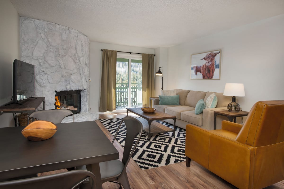Una sala de estar con pared de piedra, chimenea, sofá, un sillón amarillo y un comedor con mesa y sillas.